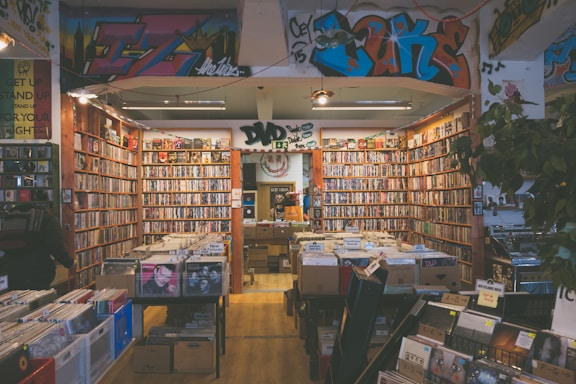A cozy music store filled with vinyl records and DVDs, featuring colorful graffiti on the walls. Shelves are lined with a vast collection of CDs and records in an organized manner. Soft lighting adds to the inviting atmosphere, and a few customers are browsing the selection.