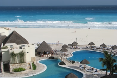 A luxurious beachfront resort features a large pool area with a winding pool bordered by stone pathways and several thatched-roof huts. Sunbeds are placed around the pool, with a few people relaxing under the sun. In the background, a white sandy beach meets a vibrant blue ocean, with waves crashing onto the shore.