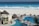 A luxurious beachfront resort features a large pool area with a winding pool bordered by stone pathways and several thatched-roof huts. Sunbeds are placed around the pool, with a few people relaxing under the sun. In the background, a white sandy beach meets a vibrant blue ocean, with waves crashing onto the shore.