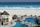 A luxurious beachfront resort features a large pool area with a winding pool bordered by stone pathways and several thatched-roof huts. Sunbeds are placed around the pool, with a few people relaxing under the sun. In the background, a white sandy beach meets a vibrant blue ocean, with waves crashing onto the shore.