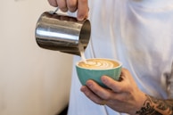 A person is skillfully pouring steamed milk from a metallic pitcher into a cup of coffee, creating a beautiful latte art pattern on the surface. The cup is turquoise and held with tattooed hands.