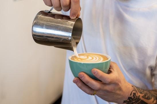 A person is skillfully pouring steamed milk from a metallic pitcher into a cup of coffee, creating a beautiful latte art pattern on the surface. The cup is turquoise and held with tattooed hands.