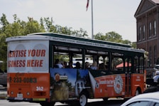 A tour bus promoting local attractions is driving along a street. The bus is mainly white, green, and orange with advertisements for local tours and a call to advertise on the vehicle. Inside, passengers can be seen seated, enjoying the ride. In the background, there are trees and a brick building with tall windows, giving a sense of an urban or historical area.