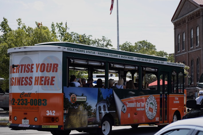 A tour bus promoting local attractions is driving along a street. The bus is mainly white, green, and orange with advertisements for local tours and a call to advertise on the vehicle. Inside, passengers can be seen seated, enjoying the ride. In the background, there are trees and a brick building with tall windows, giving a sense of an urban or historical area.