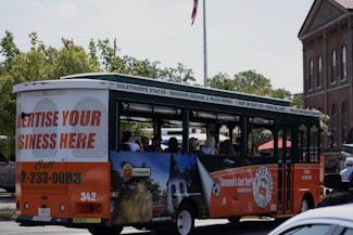 A tour bus promoting local attractions is driving along a street. The bus is mainly white, green, and orange with advertisements for local tours and a call to advertise on the vehicle. Inside, passengers can be seen seated, enjoying the ride. In the background, there are trees and a brick building with tall windows, giving a sense of an urban or historical area.