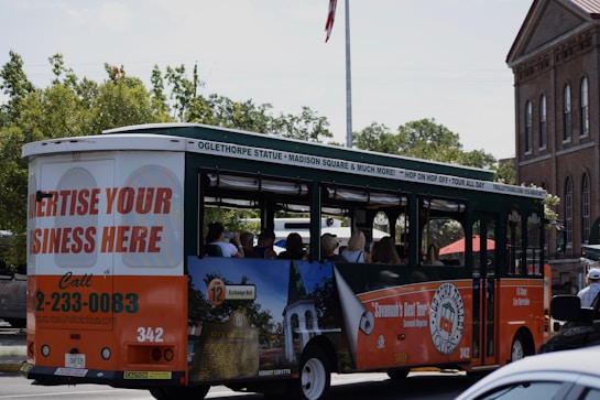 A tour bus promoting local attractions is driving along a street. The bus is mainly white, green, and orange with advertisements for local tours and a call to advertise on the vehicle. Inside, passengers can be seen seated, enjoying the ride. In the background, there are trees and a brick building with tall windows, giving a sense of an urban or historical area.