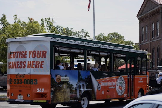 A tour bus promoting local attractions is driving along a street. The bus is mainly white, green, and orange with advertisements for local tours and a call to advertise on the vehicle. Inside, passengers can be seen seated, enjoying the ride. In the background, there are trees and a brick building with tall windows, giving a sense of an urban or historical area.