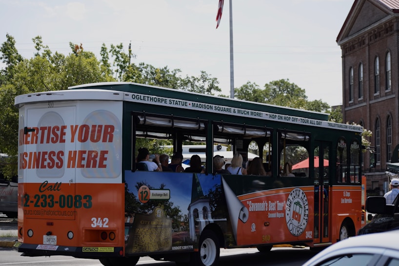 A tour bus promoting local attractions is driving along a street. The bus is mainly white, green, and orange with advertisements for local tours and a call to advertise on the vehicle. Inside, passengers can be seen seated, enjoying the ride. In the background, there are trees and a brick building with tall windows, giving a sense of an urban or historical area.