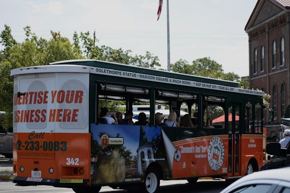 A tour bus promoting local attractions is driving along a street. The bus is mainly white, green, and orange with advertisements for local tours and a call to advertise on the vehicle. Inside, passengers can be seen seated, enjoying the ride. In the background, there are trees and a brick building with tall windows, giving a sense of an urban or historical area.
