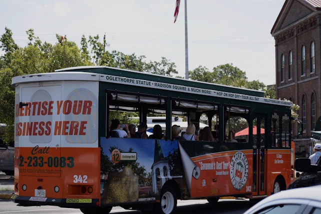A tour bus promoting local attractions is driving along a street. The bus is mainly white, green, and orange with advertisements for local tours and a call to advertise on the vehicle. Inside, passengers can be seen seated, enjoying the ride. In the background, there are trees and a brick building with tall windows, giving a sense of an urban or historical area.