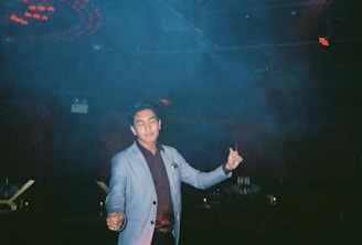 A person wearing a light-colored blazer and dark shirt is standing in an indoor setting with a relaxed expression and slightly closed eyes. The background is dimly lit with some ambient lighting and smoke or fog, suggesting a nightclub or lounge environment.