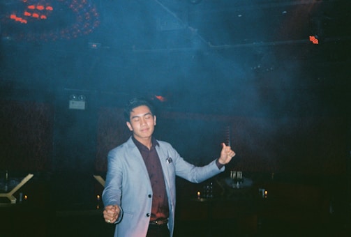 A person wearing a light-colored blazer and dark shirt is standing in an indoor setting with a relaxed expression and slightly closed eyes. The background is dimly lit with some ambient lighting and smoke or fog, suggesting a nightclub or lounge environment.