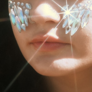 A close-up of a person's face with decorative crystal-like gems around the eyes, reflecting light and creating starburst patterns. The image focuses on the person's lips and nose, with a nose ring visible.