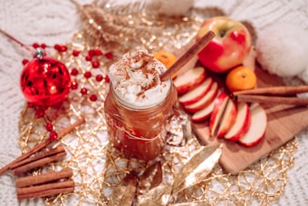A festively arranged setting contains a mug of whipped cream-topped hot beverage with cinnamon, surrounded by slices of red apple, whole apples, cinnamon sticks, a red Christmas ornament, red berries, and golden decorative elements on a cozy textured surface.