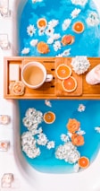 A bathtub filled with vibrant blue water, decorated with floating white and orange flowers, as well as slices of oranges. A wooden bath tray holds a white mug with a warm beverage, a bar of soap, more orange slices, and a candle.