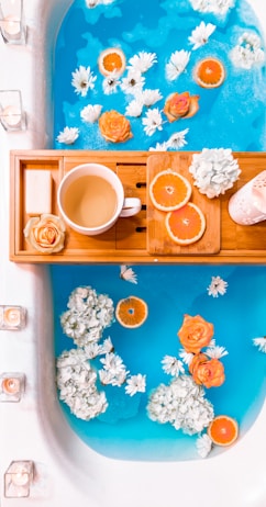 A bathtub filled with vibrant blue water, decorated with floating white and orange flowers, as well as slices of oranges. A wooden bath tray holds a white mug with a warm beverage, a bar of soap, more orange slices, and a candle.