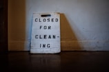 A worn-out sign made of wood rests against a wall, with the words 'CLOSED FOR CLEANING' stenciled in black capital letters. The surrounding environment appears dimly lit, suggesting an indoor setting with a rustic or aged atmosphere.