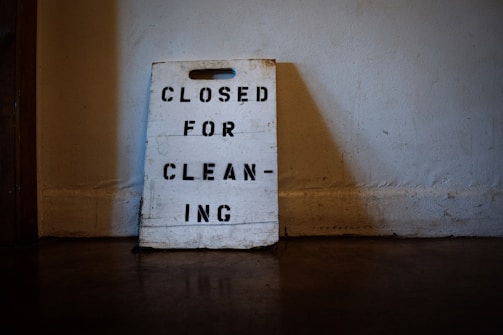 A worn-out sign made of wood rests against a wall, with the words 'CLOSED FOR CLEANING' stenciled in black capital letters. The surrounding environment appears dimly lit, suggesting an indoor setting with a rustic or aged atmosphere.