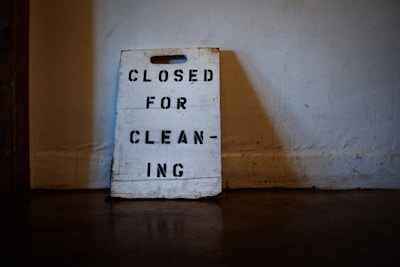 A worn-out sign made of wood rests against a wall, with the words 'CLOSED FOR CLEANING' stenciled in black capital letters. The surrounding environment appears dimly lit, suggesting an indoor setting with a rustic or aged atmosphere.
