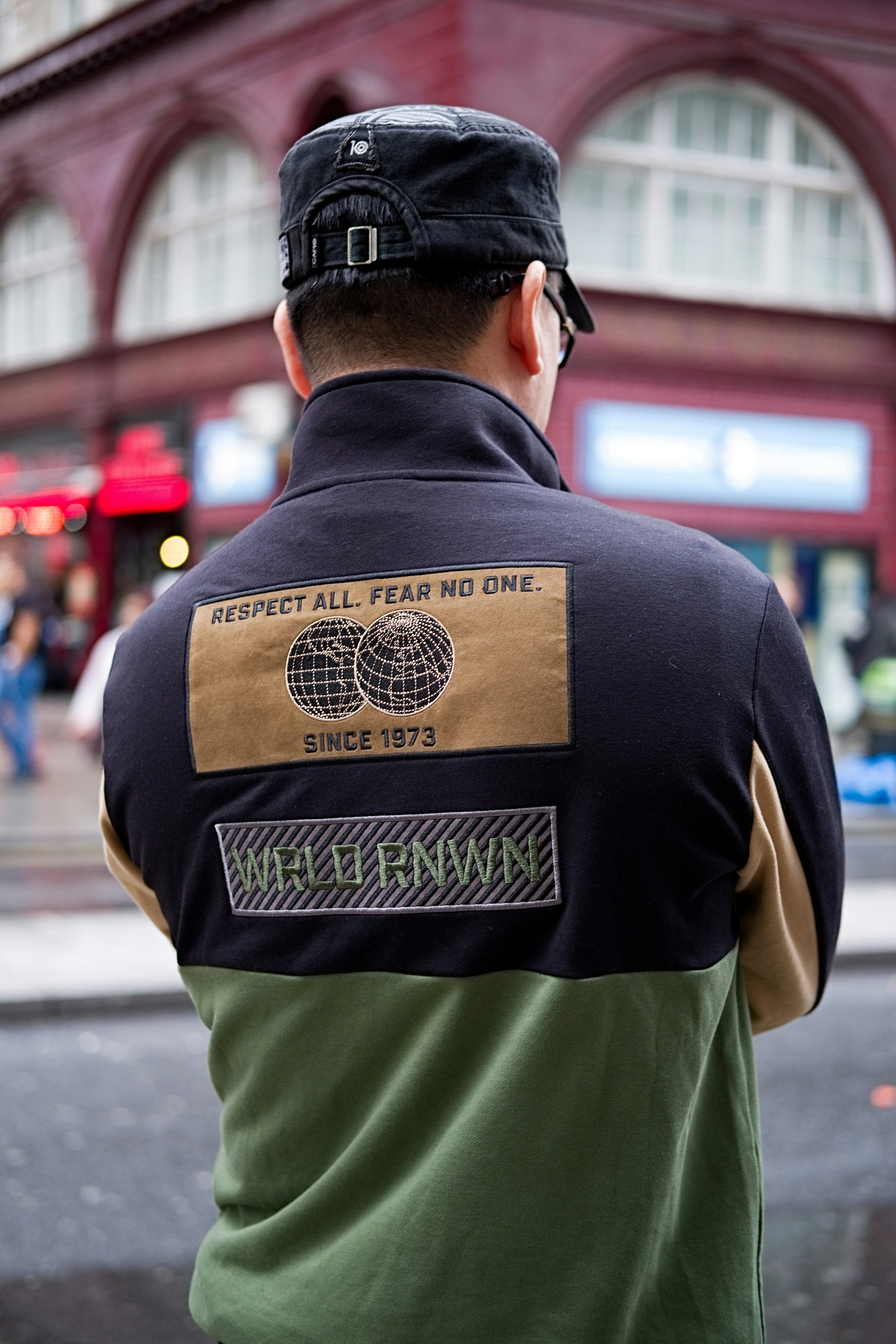 A person wearing a black cap and a color-blocked jacket with patches is standing with their back turned in an urban setting. The jacket has a design with text stating 'Respect All. Fear No One.' along with images of two globes and other text such as 'Since 1973' and 'WRLD RNWN'. The background shows a red-brick building with large windows and some signs indicating a busy street.