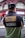 A person wearing a black cap and a color-blocked jacket with patches is standing with their back turned in an urban setting. The jacket has a design with text stating 'Respect All. Fear No One.' along with images of two globes and other text such as 'Since 1973' and 'WRLD RNWN'. The background shows a red-brick building with large windows and some signs indicating a busy street.