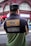 A person wearing a black cap and a color-blocked jacket with patches is standing with their back turned in an urban setting. The jacket has a design with text stating 'Respect All. Fear No One.' along with images of two globes and other text such as 'Since 1973' and 'WRLD RNWN'. The background shows a red-brick building with large windows and some signs indicating a busy street.