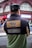 A person wearing a black cap and a color-blocked jacket with patches is standing with their back turned in an urban setting. The jacket has a design with text stating 'Respect All. Fear No One.' along with images of two globes and other text such as 'Since 1973' and 'WRLD RNWN'. The background shows a red-brick building with large windows and some signs indicating a busy street.