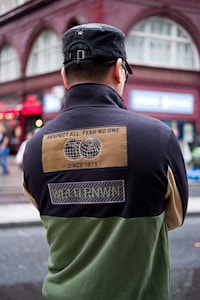 A person wearing a black cap and a color-blocked jacket with patches is standing with their back turned in an urban setting. The jacket has a design with text stating 'Respect All. Fear No One.' along with images of two globes and other text such as 'Since 1973' and 'WRLD RNWN'. The background shows a red-brick building with large windows and some signs indicating a busy street.