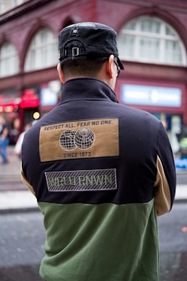 A person wearing a black cap and a color-blocked jacket with patches is standing with their back turned in an urban setting. The jacket has a design with text stating 'Respect All. Fear No One.' along with images of two globes and other text such as 'Since 1973' and 'WRLD RNWN'. The background shows a red-brick building with large windows and some signs indicating a busy street.