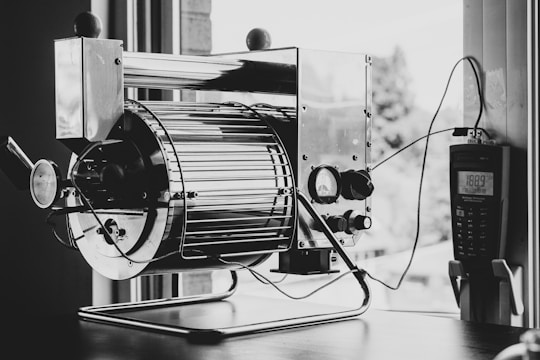 A black and white image featuring a metal roasting or industrial machine with a cylindrical drum and various control knobs. The machine is placed indoors near a window, allowing natural light to come in. To the right side of the machine, a digital thermometer or control device is visible, connected to the machine by wires. The background shows some blurred outdoor elements through the window.