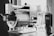A black and white image featuring a metal roasting or industrial machine with a cylindrical drum and various control knobs. The machine is placed indoors near a window, allowing natural light to come in. To the right side of the machine, a digital thermometer or control device is visible, connected to the machine by wires. The background shows some blurred outdoor elements through the window.