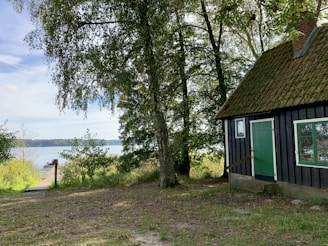 A rustic wooden cabin with a green door and moss-covered roof is nestled among tall trees. The scene overlooks a serene lake with a wooden pier extending into it. Lush greenery surrounds the area, enhancing the natural and tranquil atmosphere.