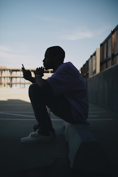A person is sitting on a concrete barrier in a contemplative pose, looking at their smartphone. The image is silhouetted against a clear sky, creating a sense of calm and introspection.
