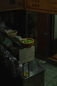 A dimly lit food stall featuring bowls of fresh green vegetables in baskets placed on a counter. There are dishes, cookware, and a menu sign with handwritten text in an Asian script. The surroundings suggest a small, possibly street-side or indoor eatery with ambiance captured in low-light conditions.