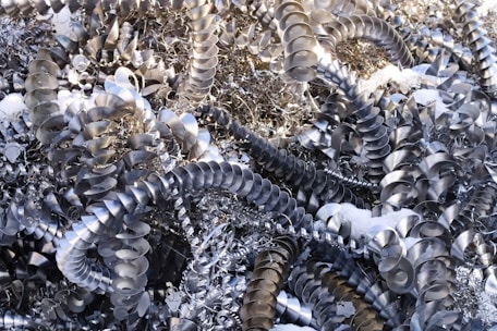 A tangled mass of metallic shavings and spirals, likely from a machining process, is shown with some patches of white snow collected among the metal. The surfaces are shiny and reflect the light, creating a contrast between the cold metallic texture and the soft snow.