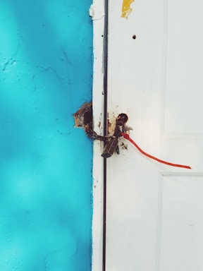 A white wooden door is secured with a rusty latch and a red plastic security seal. The door is adjacent to a bright turquoise wall, with some visible weathering and chipped paint.