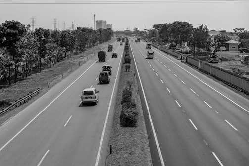 A wide highway with multiple lanes in both directions is shown. There are several vehicles, including cars and trucks, traveling along the road. Trees and utility poles line the sides of the highway. Construction activity is visible on one side, suggesting urban development.
