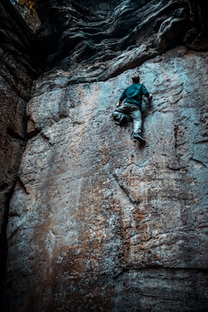 A person is climbing a rugged rock wall that features various textures and shades. The wall is steep and appears challenging to climb. The climber is dressed in casual outdoor clothing, focused and making progress upward.
