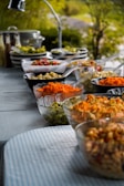 A buffet table is set outdoors with a variety of colorful salads and food items displayed in bowls. The table is covered with a blue-striped tablecloth and surrounded by greenery, creating a fresh and inviting atmosphere. Glass and ceramic bowls contain ingredients like shredded carrots, diced vegetables, and pasta, with serving utensils placed beside them.
