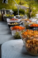 A buffet table is set outdoors with a variety of colorful salads and food items displayed in bowls. The table is covered with a blue-striped tablecloth and surrounded by greenery, creating a fresh and inviting atmosphere. Glass and ceramic bowls contain ingredients like shredded carrots, diced vegetables, and pasta, with serving utensils placed beside them.