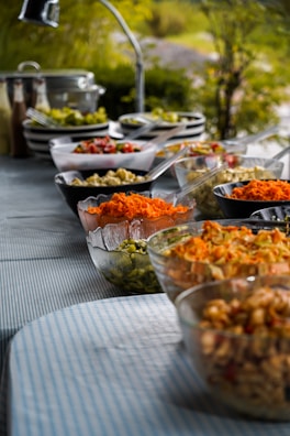 A buffet table is set outdoors with a variety of colorful salads and food items displayed in bowls. The table is covered with a blue-striped tablecloth and surrounded by greenery, creating a fresh and inviting atmosphere. Glass and ceramic bowls contain ingredients like shredded carrots, diced vegetables, and pasta, with serving utensils placed beside them.