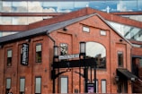 A red brick building with large arched windows and rectangular windows, featuring a sign that reads 'The Foundry'. The structure has a contemporary architectural style with a combination of brick and glass. The upper part of the building includes glass windows reflecting clouds, creating a contrast between the industrial aesthetic and modern elements.