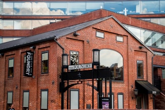 A red brick building with large arched windows and rectangular windows, featuring a sign that reads 'The Foundry'. The structure has a contemporary architectural style with a combination of brick and glass. The upper part of the building includes glass windows reflecting clouds, creating a contrast between the industrial aesthetic and modern elements.