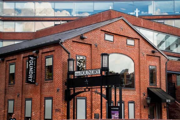 A red brick building with large arched windows and rectangular windows, featuring a sign that reads 'The Foundry'. The structure has a contemporary architectural style with a combination of brick and glass. The upper part of the building includes glass windows reflecting clouds, creating a contrast between the industrial aesthetic and modern elements.