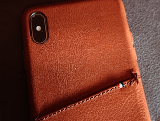 A close-up view of a smartphone with a dual camera placed inside a textured leather case, featuring detailed stitching and a small Italian flag-colored ribbon attached.