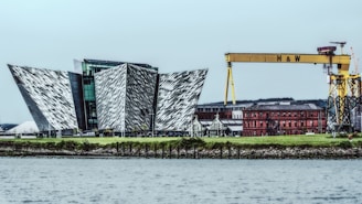 A modern architectural structure with angular, metallic surfaces is situated near the waterfront. Adjacent to the building, a large yellow gantry crane with 'H & W' written on it dominates the skyline. In the background, a red-brick industrial building adds contrast to the scene with its historical style.