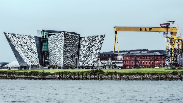 A modern architectural structure with angular, metallic surfaces is situated near the waterfront. Adjacent to the building, a large yellow gantry crane with 'H & W' written on it dominates the skyline. In the background, a red-brick industrial building adds contrast to the scene with its historical style.