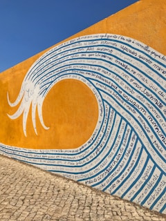 A mural painted on a wall with vibrant orange and blue hues depicting a wave. The wave is filled with handwritten text in a script style. The wall forms the background, and the surface of the ground appears to be cobblestone, adding texture to the scene.