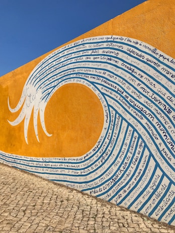 A mural painted on a wall with vibrant orange and blue hues depicting a wave. The wave is filled with handwritten text in a script style. The wall forms the background, and the surface of the ground appears to be cobblestone, adding texture to the scene.