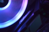 A close-up view of a computer component with a glowing LED ring in shades of blue and purple. The logo 'ASRock' is visible on the device, highlighting part of the internal hardware setup.